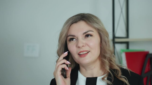 Close up of business woman with styled blond hair engaged in cheerful conversation over phone with client, showing expressive facial features, in modern office with shelf and red folder in background