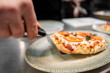Close-up of a hand lifting a hot slice of cheese pizza with tomato and herbs using a spatula,...