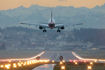 Commercial airliner landing at dusk on illuminated runway with city and mountains in background,...