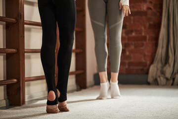 Two Caucasian teenagers practicing rhythmic gymnastics training, standing on toes in ballet position, lower bodies visible, focusing on technique and posture in gym environment