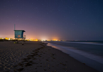 A tranquil beach scene at night, featuring a lifeguard tower and city lights on the horizon.