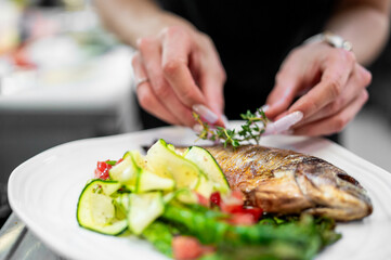 Chef garnishing grilled fish with herbs on a plate of fresh salad, zucchini, and tomatoes. Close-up of professional food styling in restaurant kitchen.