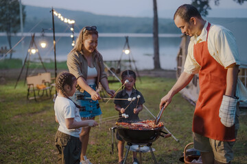 A man is cooking food on a grill while two children are making s'mores. A woman is also present, helping the children