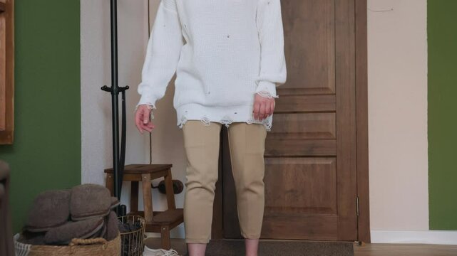 Patient stands in cozy room after removing canvas shoes, preparing to wear crocs, wearing beige pants and oversized white sweater, in front of wooden door with green walls, and indoor furniture