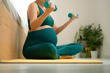 Serene pregnant female in cross-legged stretch pose on bed, maintaining fitness routine. Nurturing body and mind while awaiting baby's arrival.