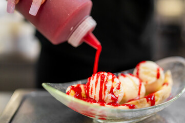 Hand pouring red syrup over scoops of vanilla ice cream in a glass dessert bowl. Close-up image...