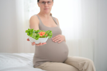 Pregnant woman balancing vegetable salad plate while gently stroking her belly. Thoughtful nutrition choice during precious maternal moments.