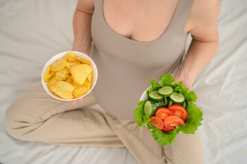 Pregnant woman holding both wholesome salad and salty chips. Authentic snapshot of dietary challenges and choices while expecting.