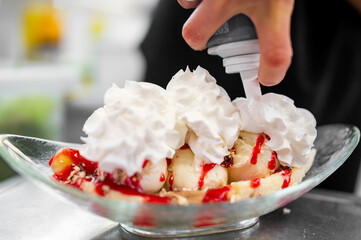 Hand adding whipped cream to a banana split dessert with vanilla ice cream, strawberry sauce, and...