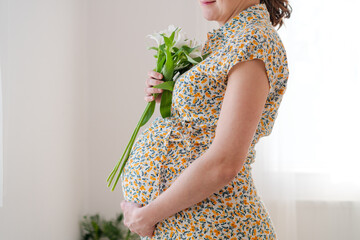 Expecting mother gently holding white flowers while touching her baby bump. Soft natural light enhances this tender maternal moment at home.
