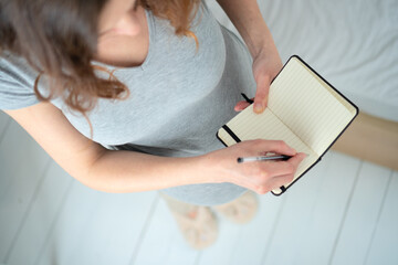 Overhead close-up of expecting mother carefully preparing birth plan in notebook. Organized maternity preparation and important reminders for upcoming delivery.