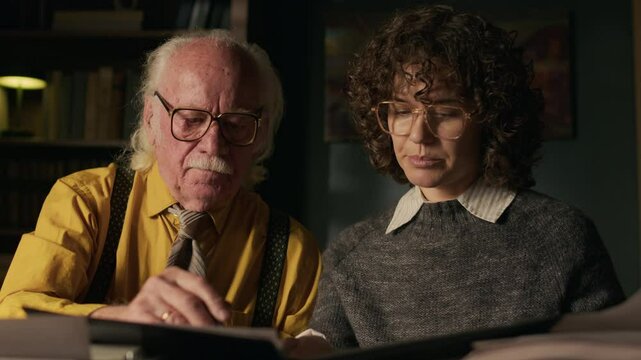Medium shot of confident middle-aged Caucasian female editor discussing manuscript with senior writer sitting at desk in his retro home study and preparing book for publishing