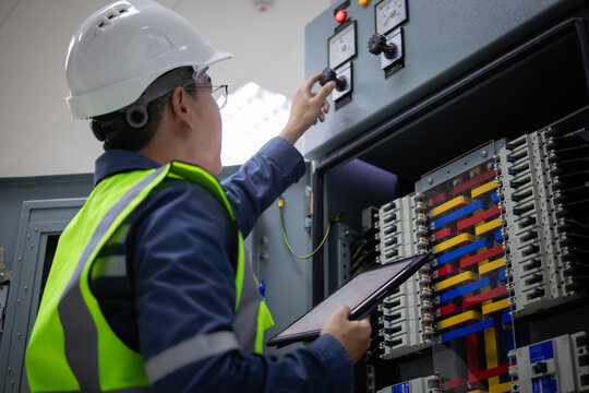 Engineer in safety vest and helmet performs inspection on electrical load center unit adjusting controls while holding tablet to ensure proper function and safety compliance