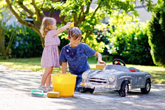 Two happy children washing big old toy car in summer garden, outdoors. Brother boy and little sister toddler girl cleaning car with soap and water, having fun with splashing and playing with sponge.
