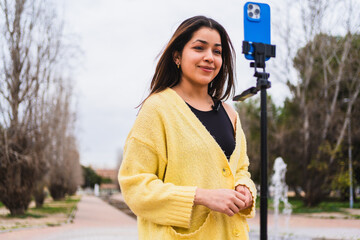 Young woman creating online content using smartphone on tripod with lavalier microphone in a park
