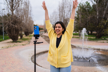 Young woman vlogger recording video for social media with smartphone on tripod, waving to her followers in urban park