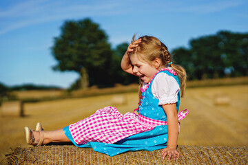 Cute little kid girl in traditional Bavarian costume in wheat field. Happy child with hay bale...