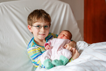 Happy boy with glasses holding his sleeping newborn sister in hospital bed. Real life, real people...