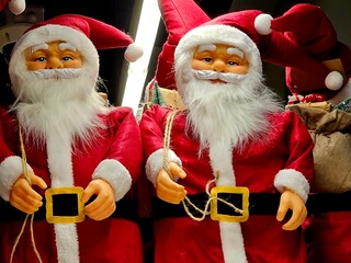Two almost identical Santa Claus figurines with white beards, red suits, and hats stand side by...