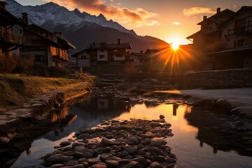 Sun setting behind a mountain village, reflecting on a river with rocks in the foreground