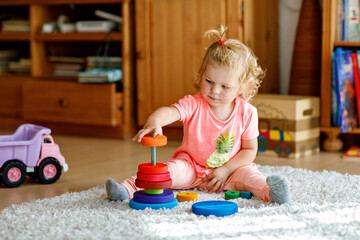 Happy toddler girl sits on a carpet, playing with colorful wooden rings and a toy truck, focused on stacking the pieces. Child plays with rainbow colored pyramid at home or nursery.