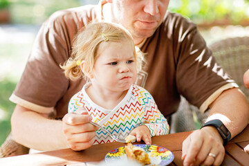 Young middle-aged father feeding cute little toddler girl in restaurant. Kid boy eating on background. Happy healthy family in an outdoor cafe in summer time, eating cake