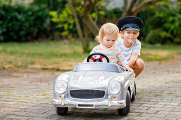 Two happy children playing with big old toy car in summer garden, outdoors. Kid boy driving car with little toddler girl inside. Laughing and smiling kids. Family, childhood, lifestyle concept