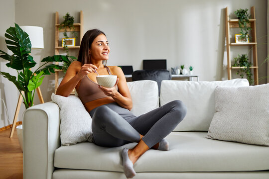 Young athletic woman eating healthy breakfast at home sitting on sofa