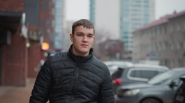 Young automobile enthusiast in black quilted winter coat walking along urban pavement past row of parked vehicles beside brick buildings with blurred city background on overcast gray day