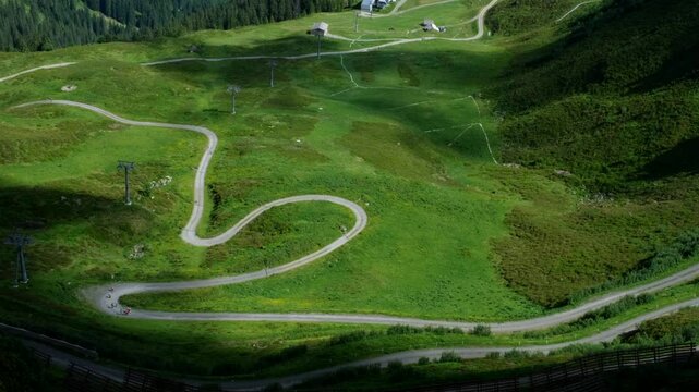 Winding mountain road cuts through lush green hills with scattered bikers riding along the trail. Overhead ski lifts and alpine meadows complete the scenic view.