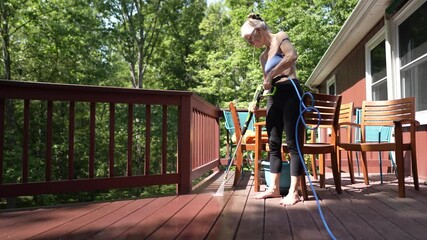 A mature woman is using a pressure washer to clean her wooden deck, ensuring it is free of dirt and grime while working in a bright garden setting.