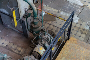 Top view of an old industrial installation with valves, pipes, and a pressure gauge, located next to a metal railing and stairs. Technical interior showing clear signs of wear and use