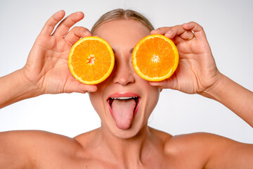Young woman playfully holds orange slices in front of her eyes with her tongue sticking out,...