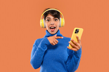 Young woman with headphones enjoys music on smartphone in bright orange background, expressing...