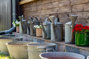 collection of old metal watering cans and galvanized tubs arranged by a wooden wall. Potted flowers...