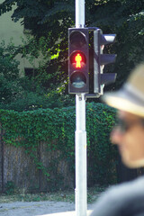 Red pedestrian traffic light glowing on summer day with blurred person in sunglasses and straw hat...
