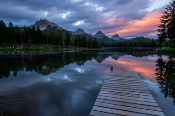 Photo of tranquil mountain lake at sunrise, fiery sky reflected on still water, jagged peaks, wooden dock with �Serenity Now� carving, evergreen silhouettes.