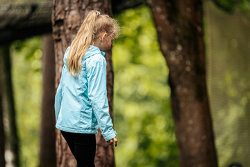 A young girl in a light blue jacket and black pants stands outdoors among tall trees, looking...