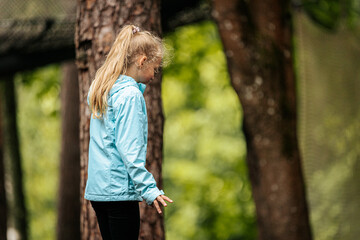 A young girl in a light blue jacket and black pants stands outdoors among tall trees, looking...
