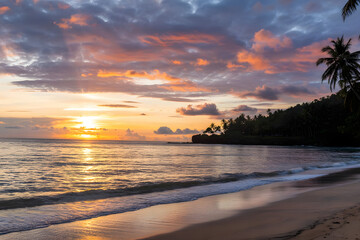 Tropical beach sunrise with orange, pink, and blue sky, calm ocean reflecting colors, palm trees, golden sand, rocks, and distant building.