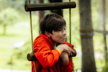 A young boy in a red jacket leans on a wooden rope ladder outdoors, looking thoughtful. Blurred...