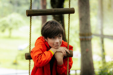 A young boy in a red jacket leans on a wooden rope ladder outdoors, looking thoughtful. Blurred...