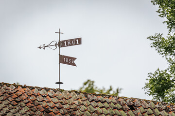 A rusty weather vane with dates 1861 and 1857 stands atop a mossy tiled roof, framed by overcast...