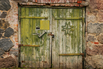 A rustic green wooden barn door with weathered paint, metal reinforcements, and a decorative bottle cap cross, set in a stone and brick wall.