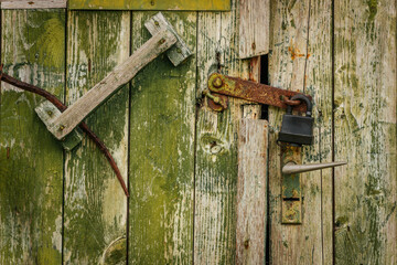 Close-up of an old green wooden door secured with a rusted latch and black padlock, showing cracked...