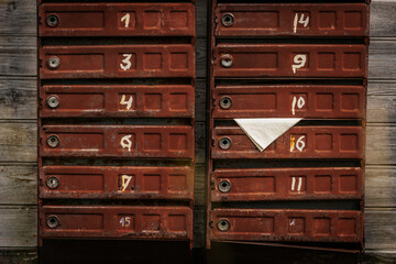 Two rows of old rusty brown mailboxes mounted on a weathered wooden wall, with handwritten white...