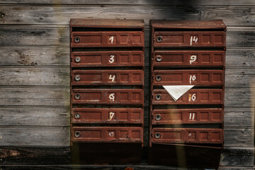 Two rows of old rusty brown mailboxes mounted on a weathered wooden wall, with handwritten white...
