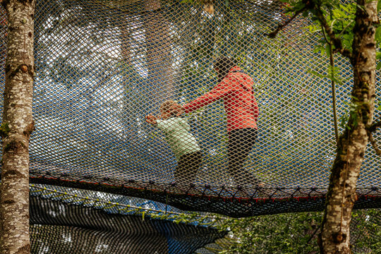 An adult and child walk together on a large safety net suspended among trees in a forest adventure park, creating a fun and active bonding moment. - Powered by Adobe