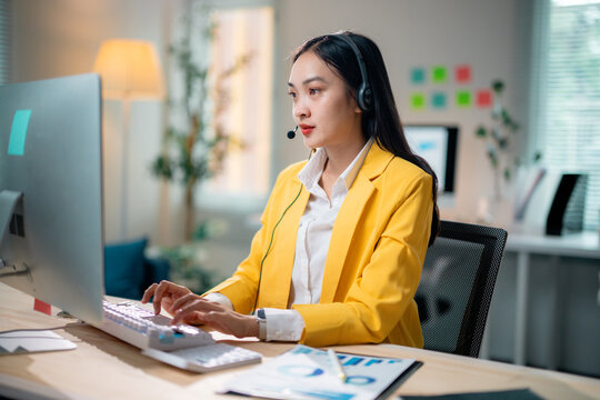 Young Asian businesswoman wearing headset is typing on keyboard and looking at computer monitor in office