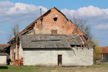 Old rural barn with exposed red brick, broken asbestos-cement roof, and patchwork repairs stands...
